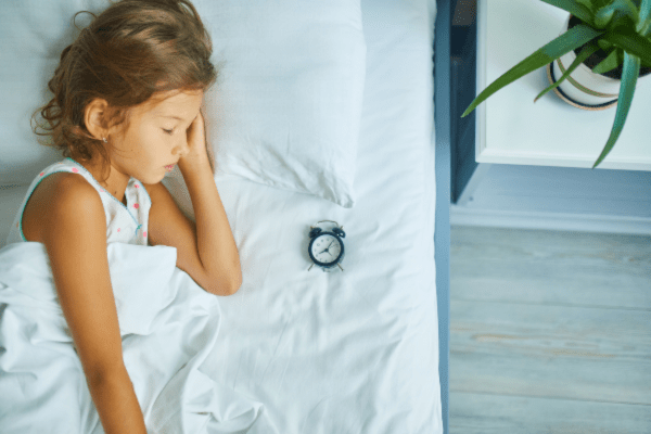 Photo of a young girl lying on her side in a bed with white sheets. Illustrating melatonin overdose in children.