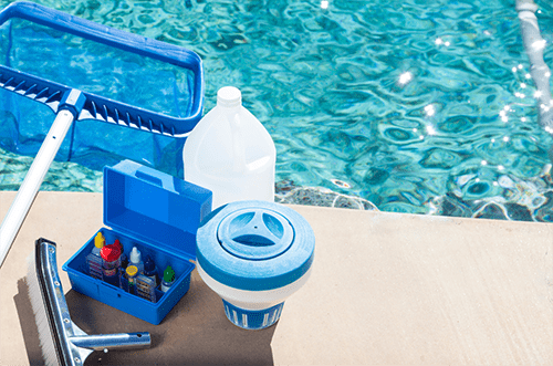 Photo of bottles of pool chemicals and equipment next to sparkling clear pool water. Depicting the safe handling and storage of pool chemicals to prevent accidents and injuries around swimming pools.